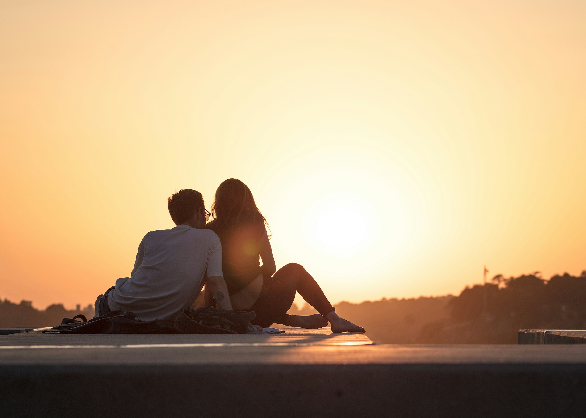 O casal sentado lado a lado, de costas, diante do pôr do sol