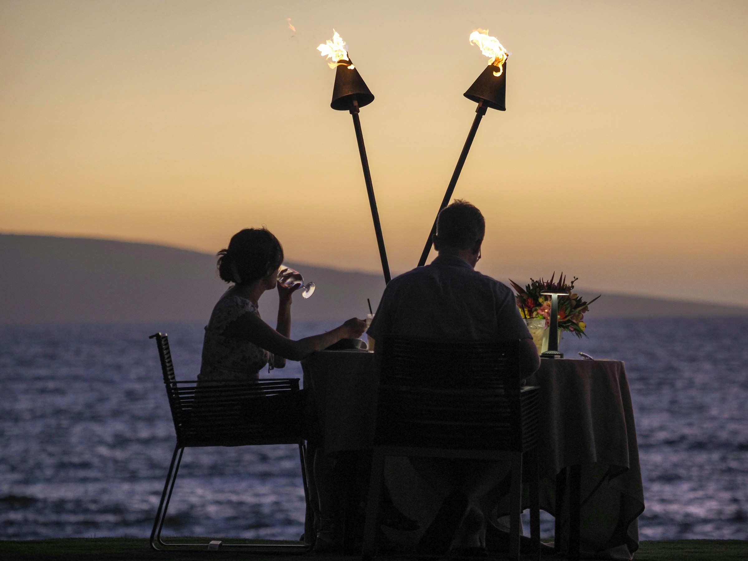 O casal jantando à beira-mar ao pôr do sol, entre tochas acesas